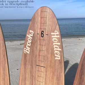 May include: Wooden growth chart rulers with names and height markings. The rulers are made of light brown wood and have white text. The background shows a beach with the ocean and sky.