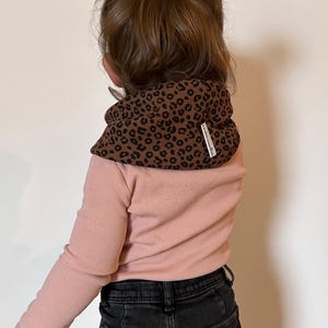 Könnte beinhalten: Ein braun-schwarzer Nackenwärmer mit Leopardenmuster, gerippter Textur und einem kleinen weißen Markenetikett. Der Nackenwärmer wird über einem langärmeligen, hellrosa gerippten Oberteil und einer dunkelgrauen Jeans getragen, dazu ein passendes Accessoire.