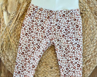 May include: White baby leggings with a brown leopard print. The leggings have a wide white waistband.
