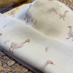 May include: Cream-colored ribbed knit beanie featuring a repeating pattern of geese in soft brown and peach tones. The hat is folded over, showcasing the interior fabric and design. The fabric has a subtle texture.
