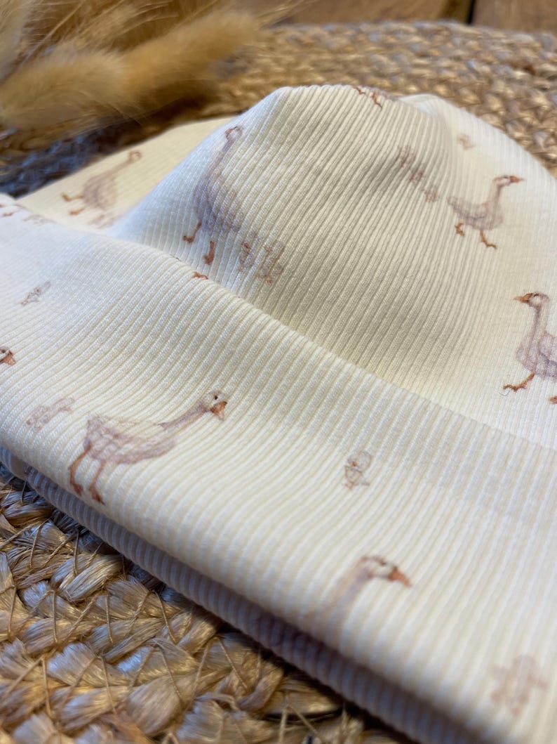 May include: Cream-colored ribbed knit beanie featuring a repeating pattern of geese in soft brown and peach tones. The hat is folded over, showcasing the interior fabric and design. The fabric has a subtle texture.