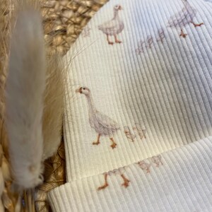 May include: A cream-colored ribbed knit beanie featuring a repeating pattern of watercolor geese. The geese are depicted in shades of gray and brown. The hat is made of a soft, ribbed fabric.