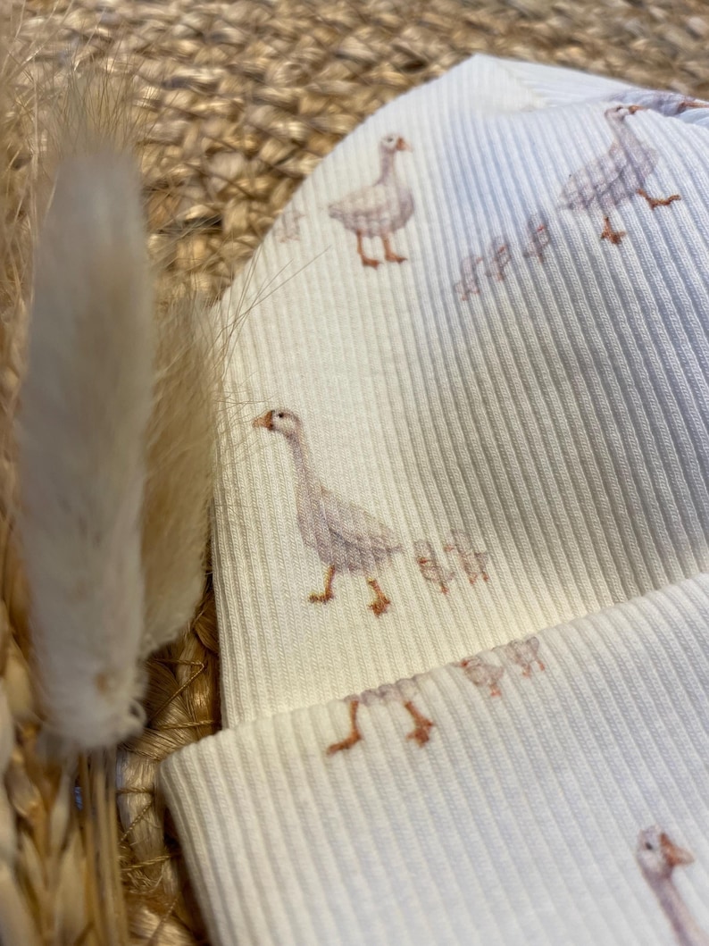 May include: A cream-colored ribbed knit beanie featuring a repeating pattern of watercolor geese. The geese are depicted in shades of gray and brown. The hat is made of a soft, ribbed fabric.