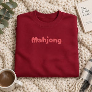 May include: A dark red crewneck sweatshirt featuring the word Mahjong embroidered in a lighter red, rounded font across the center of the chest. The garment has long sleeves and a ribbed neckline, providing a simple and clean appearance.