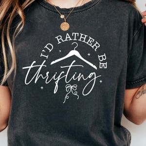 Peut inclure: T-shirt gris foncé avec l'inscription blanche "I'd Rather Be Thrifting". Le design comprend un cintre et des éléments décoratifs. Un t-shirt décontracté pour les amateurs de friperie et de shopping d'occasion.