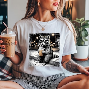 May include: White t-shirt featuring a black and white graphic of a raccoon wearing sunglasses and drinking from a cup with a straw. The raccoon is sitting on a curb. The shirt is a casual, everyday wear item.