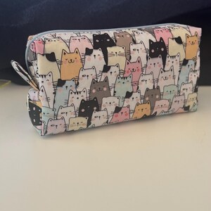 Puede incluir: Una bolsa blanca con cremallera con un patrón repetitivo de gatos de dibujos animados de colores. Los gatos están en varias poses y tienen diferentes expresiones faciales.