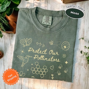 May include: A green t-shirt with the text "Protect Our Pollinators" in gold lettering. The shirt features a hummingbird, a butterfly, bees, and honeycombs.