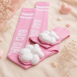 May include: Pink socks with white paw pads and the text "BAD GIRL". The socks have white stripes at the top. The socks are displayed on a soft, light-colored surface with decorative elements.