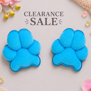 May include: Two bright blue plush paw-shaped items with soft, textured surfaces. The text CLEARANCE SALE is printed in dark gray letters above the two paw shapes. The items are arranged symmetrically on a light-colored background.