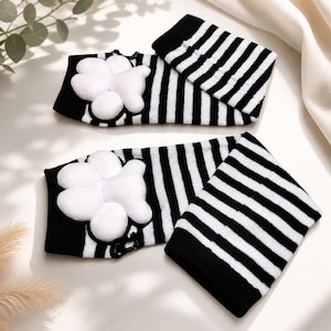 May include: Black and white striped baby leg warmers with a white paw print design. The leg warmers are made of a soft material and are designed to keep baby's legs warm. The paw print is a cute and playful detail.