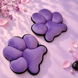 May include: Two plush, purple, paw-shaped cushions with soft, rounded toe pads and a black border trim. The items feature a textured, velvety surface and are arranged on a light purple surface alongside small pink flower petals and beads.