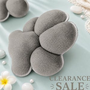 May include: A plush, grey, paw-shaped cushion with a soft, fuzzy texture and stitched edges. The item features a central pad and four rounded toe sections, mimicking the appearance of an animal paw, presented in a clean, simple design.