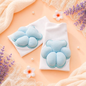 May include: Two white ribbed socks with light blue paw-shaped appliqués. The appliqués have a light blue outline. The socks are displayed on a peach-colored surface with decorative flowers and lavender sprigs.
