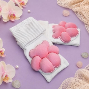 May include: A pair of white socks featuring a plush, three-dimensional pink bear paw design on the front. The socks have a ribbed cuff at the top and a soft, textured fabric construction. The bear paw applique is made of a smooth, velvet-like material.