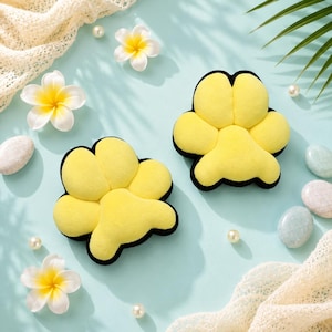 May include: Two yellow, flower-shaped plush pillows with black trim are displayed on a light blue surface. The pillows are surrounded by white and yellow flowers, pearls, and decorative stones. A white lace fabric and a green leaf add to the aesthetic.