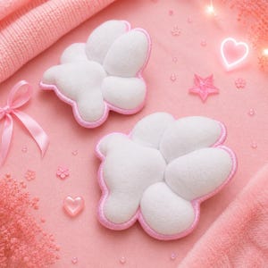 May include: Two white cloud-shaped plush pillows with pink trim are displayed on a pink surface. A pink bow, a pink heart, and a pink star are also visible. The background includes a pink knitted fabric and small decorative elements.