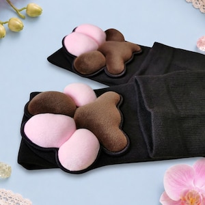 May include: A pair of black socks featuring plush, three-dimensional animal paw pads attached to the top. The paw pads are made of soft, brown and light pink fabric, designed to mimic the appearance of a cat or dog paw with distinct toe beans.