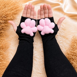 May include: Black fingerless gloves with pink paw pads. The gloves are long-sleeved and made of a soft material. The paw pads are a light pink color and are sewn onto the back of the hand.
