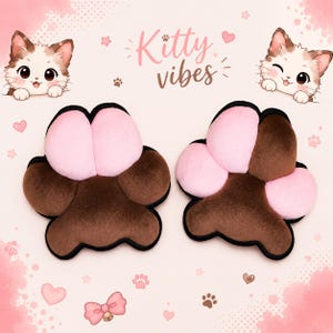 May include: Two plush cat paw-shaped cushions in brown and pink, with black trim. The image includes cartoon cat illustrations and the text "Kitty vibes" in pink. The background is light pink with paw prints and heart shapes.