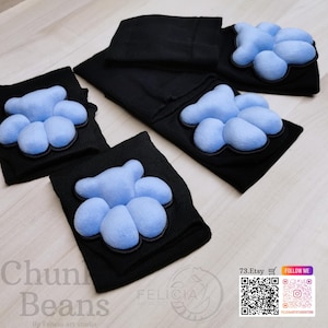 May include: Black leg warmers with light blue paw-shaped accents. The image shows several pairs of leg warmers, some folded and some unfolded, with the paw pads prominently displayed. The text "Chun Beans" is visible in the lower left corner.