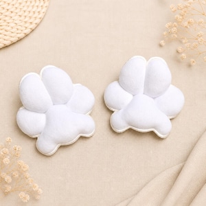 May include: Two white, paw-shaped felt decorations. Each paw has four rounded lobes and a smaller lobe at the bottom. The paws are on a beige fabric surface, with dried flowers and a woven mat in the background.