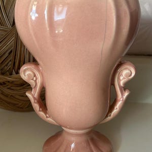 May include: A vintage pink ceramic vase with a flared top and ornate handles. The vase has a pedestal base and a glossy finish. The design is reminiscent of mid-century modern decor. The vase is approximately 7 inches tall.