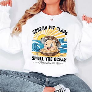 Peut inclure: Sweat-shirt blanc avec un dessin d'une huître souriante sur une plage, avec le texte "SPREAD MY FLAPS SMELL THE OCEAN Tropic Like It's Hot". Le motif comprend un soleil, des vagues et du sable.