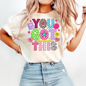 以下が含まれることがあります： クリーム色のTシャツに、カラフルで遊び心のある文字で「YOU GOT THIS」と書かれています。デザインには、ノート、リンゴ、星、デイジー、鉛筆が含まれています。文字には、ヒョウ柄やストライプなど、さまざまなパターンがあります。