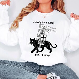 May include: White sweatshirt with a black and white graphic. The graphic features a knight on a black panther holding a flag that reads "Defend Your Local" above "public library".