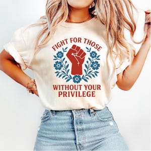 May include: Cream-colored t-shirt featuring the text "FIGHT FOR THOSE WITHOUT YOUR PRIVILEGE" in red and white. The design includes a red fist inside a blue floral wreath. The shirt is made of a soft material.