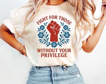 Könnte beinhalten: Cremefarbenes T-Shirt mit dem Text "FIGHT FOR THOSE WITHOUT YOUR PRIVILEGE" in Rot und Weiß. Das Design zeigt eine rote Faust in einem blauen Blumenkranz. Das Shirt ist aus einem weichen Material.