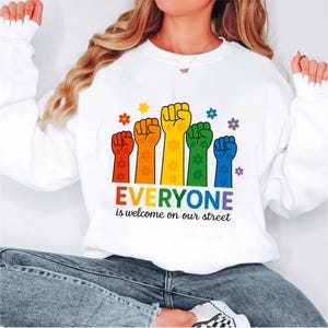 Op de afbeelding: Witte sweatshirt met een regenboog vuistafbeelding en de tekst "EVERYONE is welcome on our street". De afbeelding toont vuisten in rood, oranje, geel, groen en blauw, met bloemaccenten.