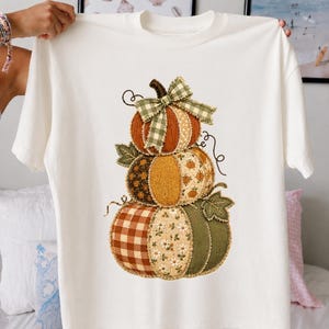 May include: A white short-sleeve t-shirt featuring a central graphic of three stacked pumpkins. The pumpkins are decorated with various fabric patterns including brown gingham, floral prints, and solid textures, topped with a green plaid fabric bow.