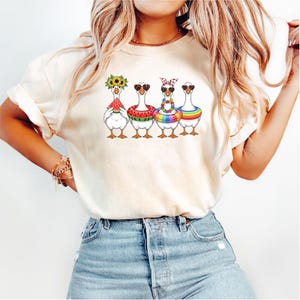May include: Cream-colored t-shirt featuring a whimsical illustration of four geese, each adorned with summer accessories. One goose wears a sunflower crown, another a watermelon slice, and the others sport sunglasses and colorful accessories. The geese are arranged in a row.