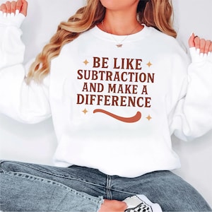 May include: White sweatshirt featuring the text "BE LIKE SUBTRACTION AND MAKE A DIFFERENCE" in brown. The design includes a curved line and starburst accents. A casual style sweatshirt.