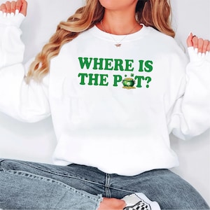 May include: White sweatshirt with the text "WHERE IS THE POT?" in green, along with a graphic of a pot of gold and a shamrock. The sweatshirt is a casual style, suitable for St. Patrick's Day.