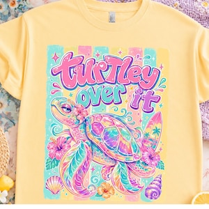 May include: Yellow t-shirt featuring a colorful graphic of a sea turtle with the text "Turtle-y over it." The turtle is adorned with flowers and a surfboard, set against a striped background. The design incorporates pastel colors.