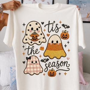 Könnte beinhalten: Ein cremefarbenes T-Shirt mit einem Grafikdruck von drei Geistern, zwei Kürbissen, einem Zuckermais und zwei Fledermäusen. Der Text lautet 'tis the season' in einer schwarzen Schreibschrift. Die Geister haben Punkte-, Streifen- und Karomuster.