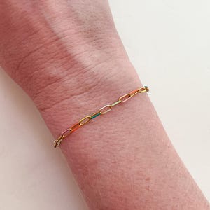 May include: A gold-colored chain bracelet with colorful enamel-filled links. The bracelet is worn on a wrist and features links in shades of pink, orange, yellow, green, and blue. The bracelet is a delicate piece of jewelry.