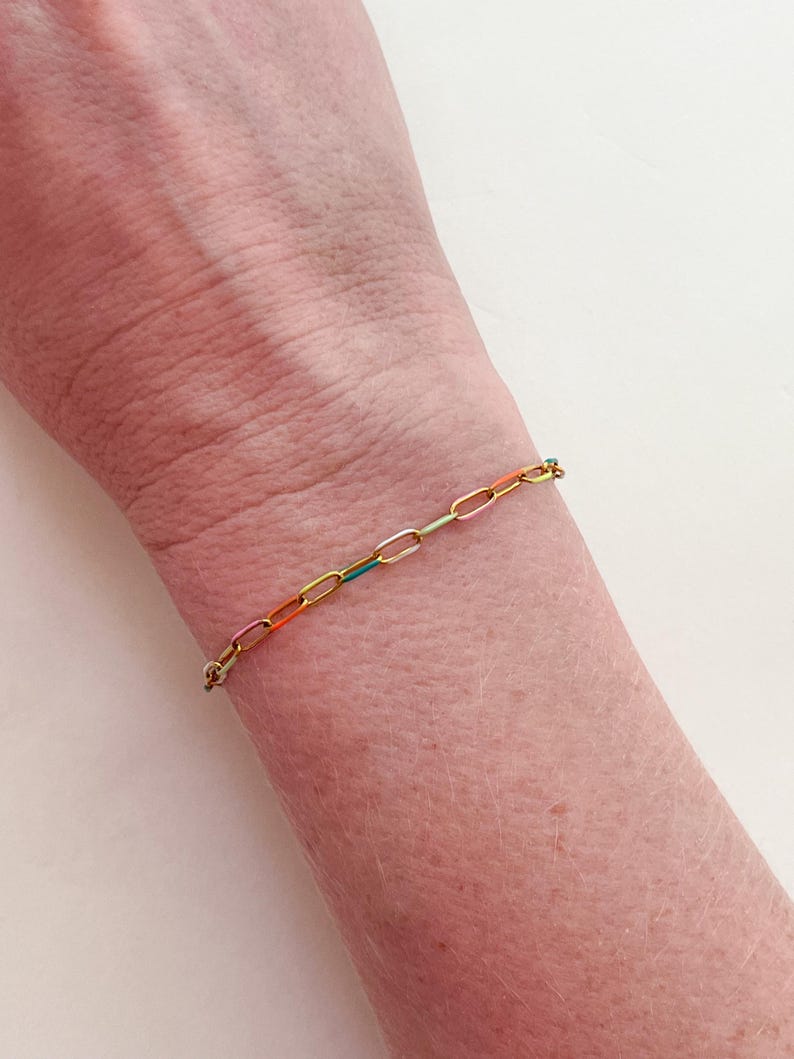 May include: A gold-colored chain bracelet with colorful enamel-filled links. The bracelet is worn on a wrist and features links in shades of pink, orange, yellow, green, and blue. The bracelet is a delicate piece of jewelry.