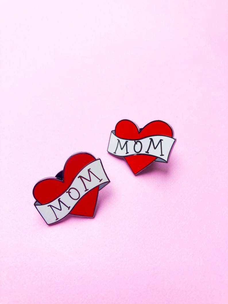 May include: Two red heart-shaped pins with white banners that say "MOM" on a pink background.