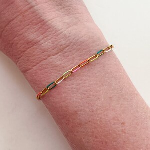 May include: A gold-colored chain bracelet with colorful enamel links. The bracelet features links in yellow, green, white, pink, and orange. The bracelet is worn on a wrist.
