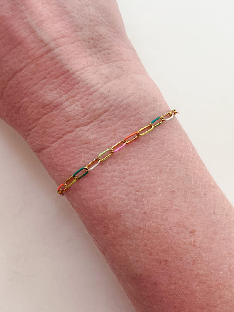 May include: A gold-colored chain bracelet with colorful enamel links. The bracelet features links in yellow, green, white, pink, and orange. The bracelet is worn on a wrist.