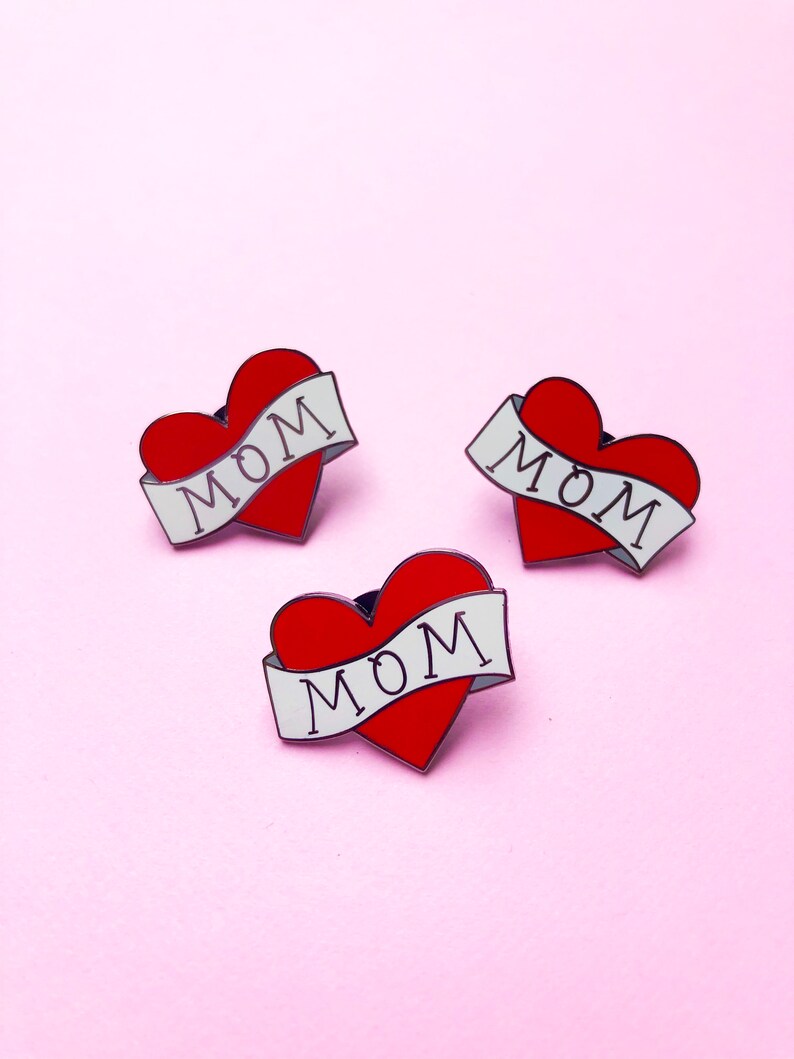 May include: Three red heart-shaped pins with white banners that say "MOM" on a pink background.