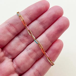 May include: A gold-colored chain bracelet with colorful enamel links in shades of teal, orange, pink, and yellow. The bracelet is held in a person's hand against a neutral background. The links are oval-shaped.