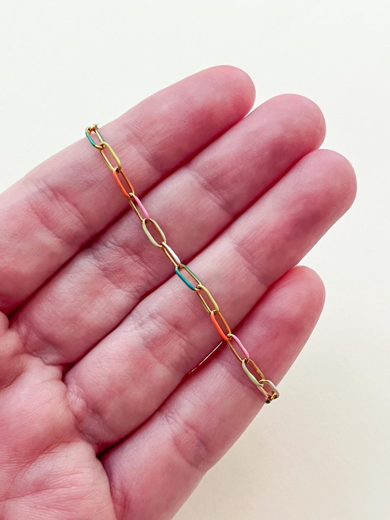 May include: A gold-colored chain bracelet with colorful enamel links in shades of teal, orange, pink, and yellow. The bracelet is held in a person's hand against a neutral background. The links are oval-shaped.