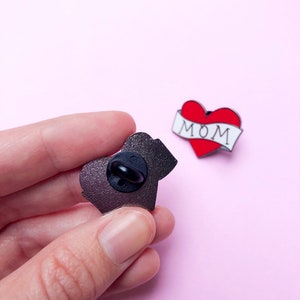 May include: A red heart with a white banner that says "MOM" on it. The back of the pin is black with a metal pin back.