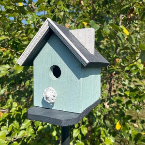 May include: A blue birdhouse with a black roof and a white decorative accent. The birdhouse is mounted on a black metal pole.