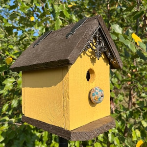 May include: A yellow birdhouse with a brown roof and a floral knob. The birdhouse is mounted on a black metal pole.